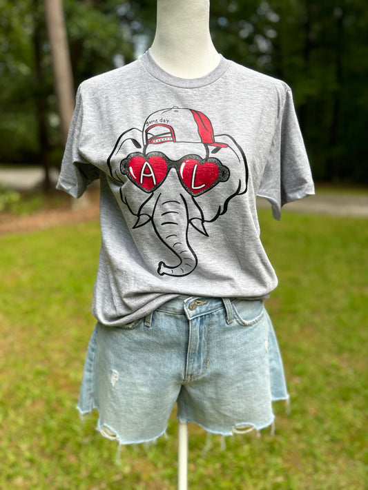 Gameday t-shirt with an Alabama elephant wearing crimson  heart shaped glasses with an A and a L in the shades and a backwards baseball hat. Stay comfortable and stylish while supporting your sports team. 