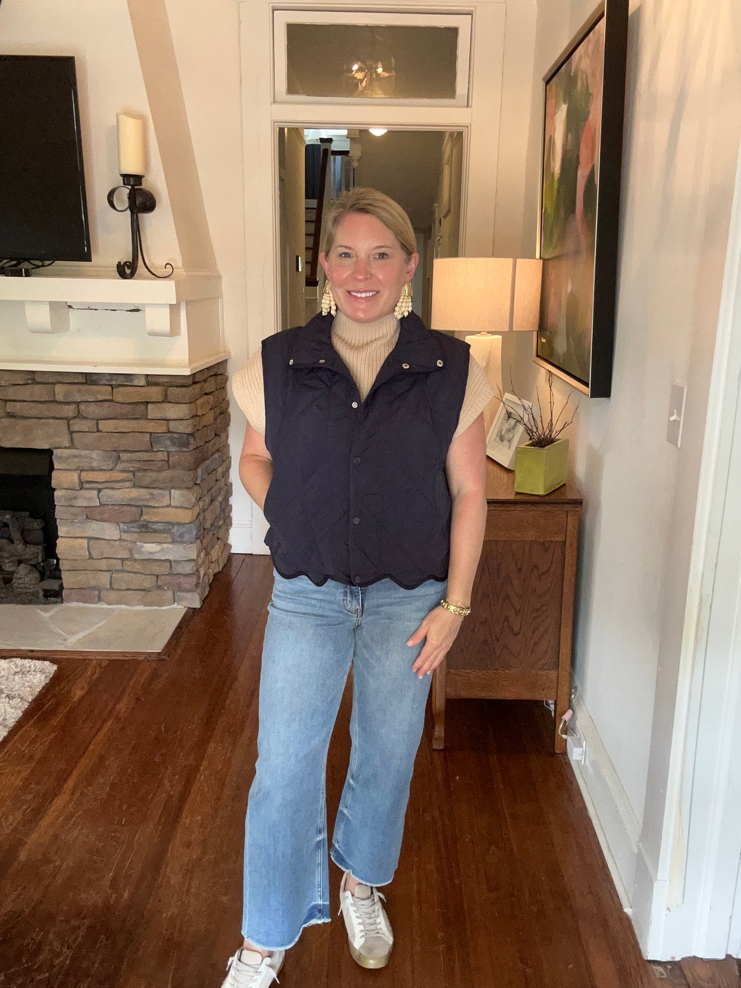 A woman standing in a home environment wearing a navy quilted snap button up vest with pockets at the side and a scalloped hem, paired with light blue jeans.