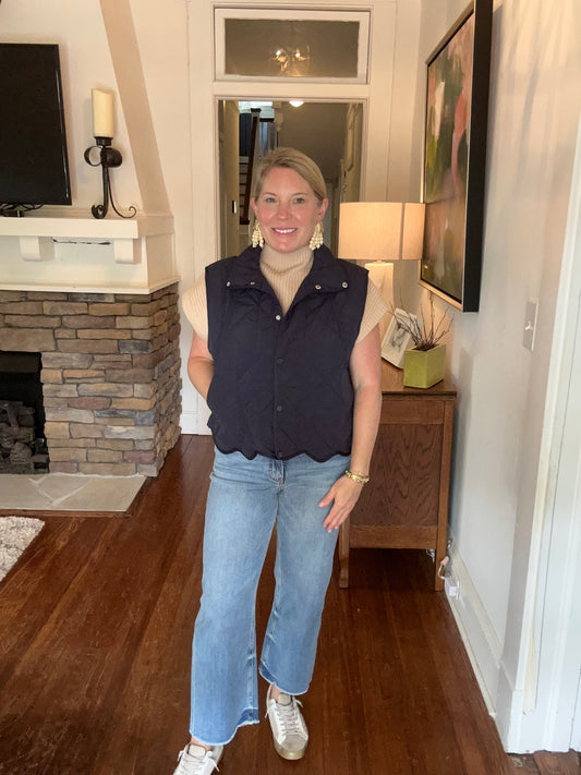 A woman standing in a home environment wearing a navy quilted snap button up vest with pockets at the side and a scalloped hem, paired with light blue jeans.